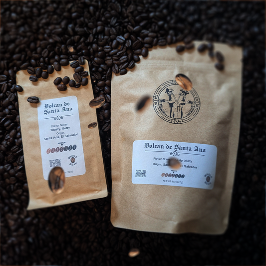 Two bags of Volcan De Santa Ana coffee, one with a label showing the product name and the other with a label showing the coffee beans and name. The bags are placed on a background of loose coffee beans scattered on a surface.